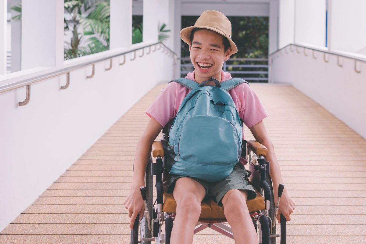 A smiling teenager in a wheelchair with a backpack and sun hat rolls down a ramp.